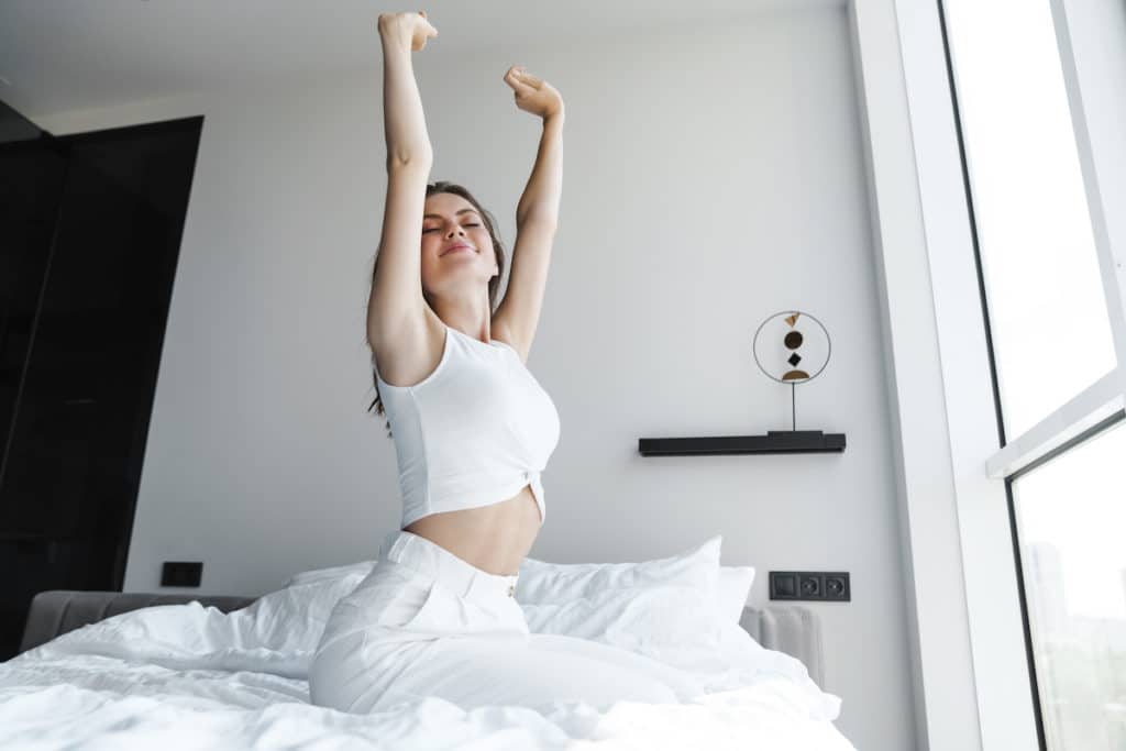 Image of young pleased woman stretching her body while sitting on bed Mommy Makeover in Southlake, TX